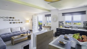 A modern living room with a sofa, coffee table, and a dining area with a fruit bowl and cups. - Talos Luxury Suites (Chania)