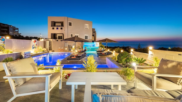 A modern outdoor pool area with lounge chairs and umbrellas, a well-lit pool, and a view of the sea at dusk. - Talos Luxury Suites (Chania)