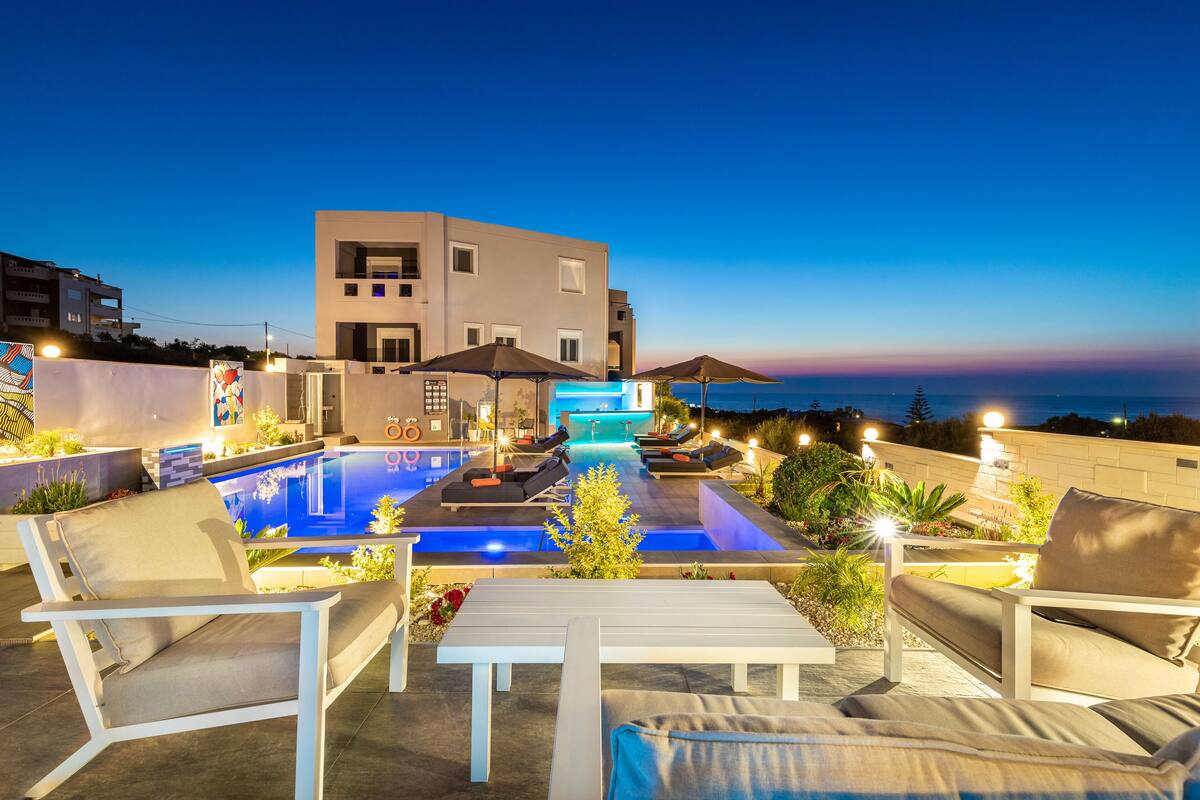 A modern outdoor pool area with lounge chairs and umbrellas, a well-lit pool, and a view of the sea at dusk.