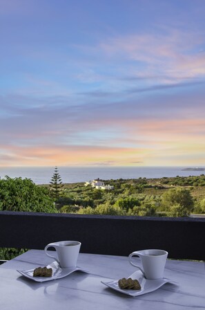 A table with two white cups and a saucer, each with a small plate of cookies, set against a scenic view of the sea and greenery during sunset. - Talos Luxury Suites (Chania)