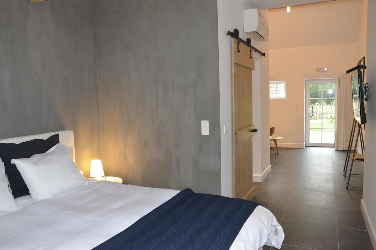 A hotel room with a bed, a nightstand, a lamp, a wooden door, and a view of the outside.
