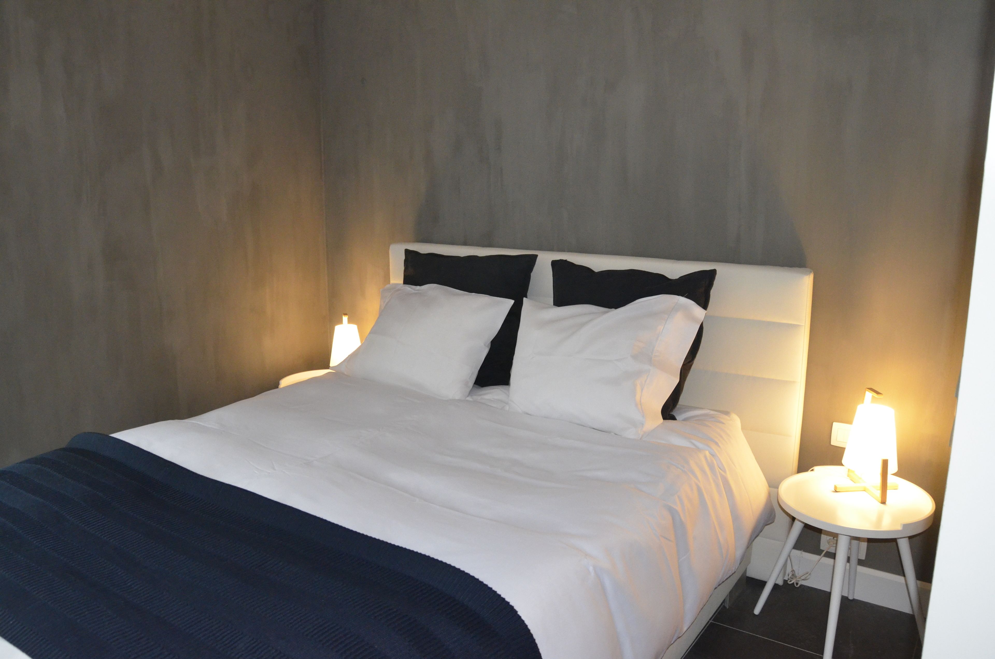 Een netjes opgemaakt bed met witte beddengoed en een donkerblauwe plaid, een nachtkastje met een lamp, en een gestructureerde wand op de achtergrond.