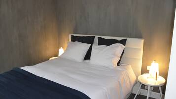 Een netjes opgemaakt bed met witte beddengoed en een donkerblauwe plaid, een nachtkastje met een lamp, en een gestructureerde wand op de achtergrond.