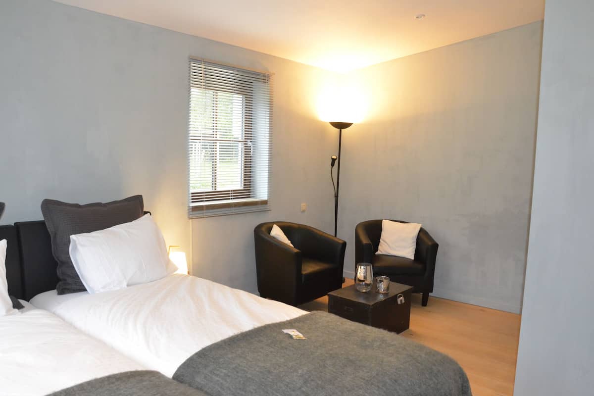 A modern hotel room with a bed, two armchairs, a bedside table, and a floor lamp.