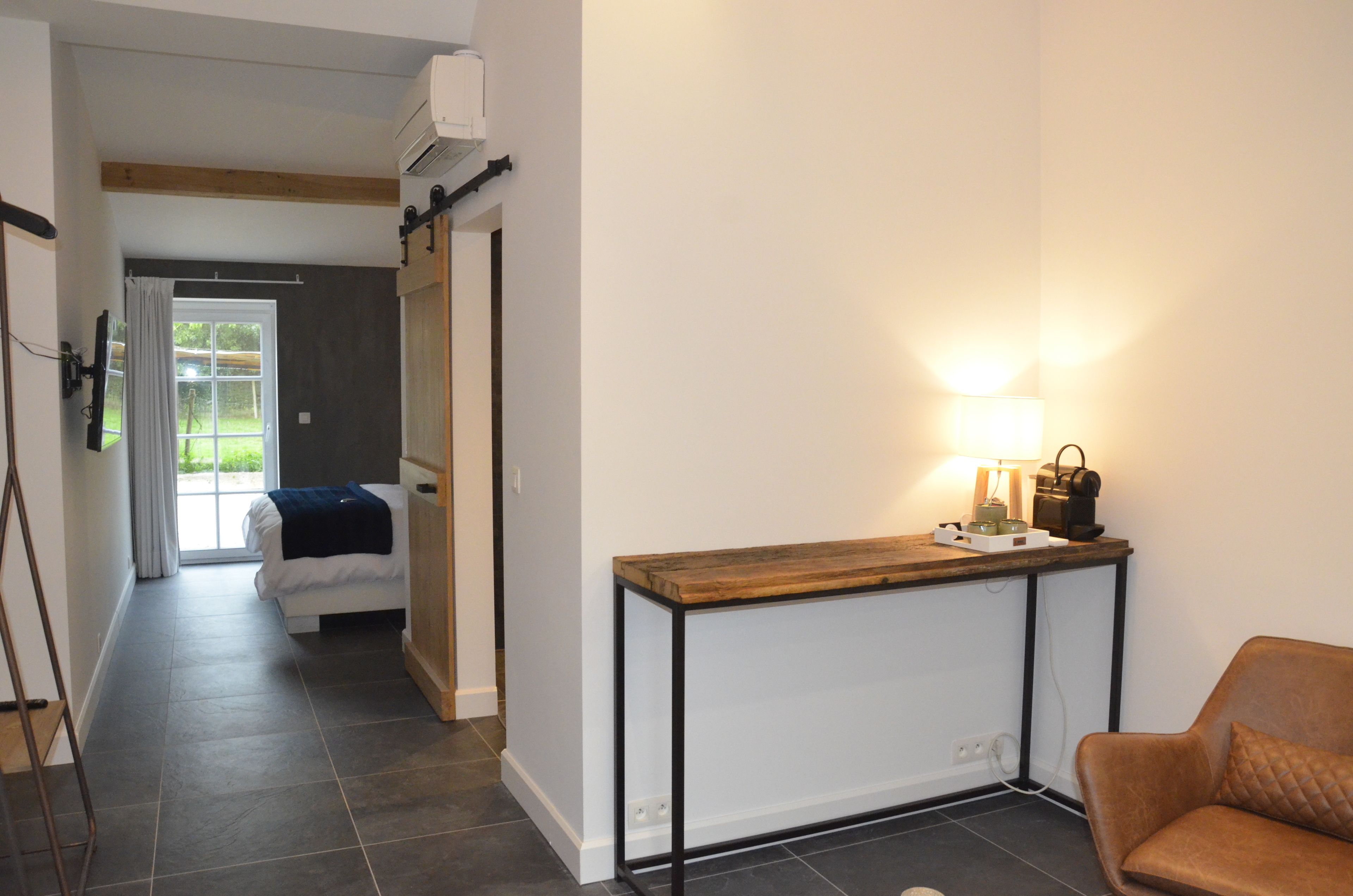 A hotel room with a bed, a bedside table with a lamp, a brown leather chair, and a view of a garden through a window.