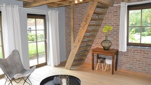 Une pièce avec un mur en briques, un escalier en bois, une chaise grise, une petite table sur laquelle est posé un vase de fleurs, et une fenêtre avec des rideaux.