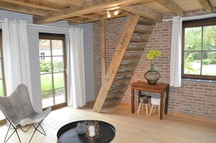Una habitación con pared de ladrillo, escalera de madera, una silla gris, una mesita con un florero y una ventana con cortinas.