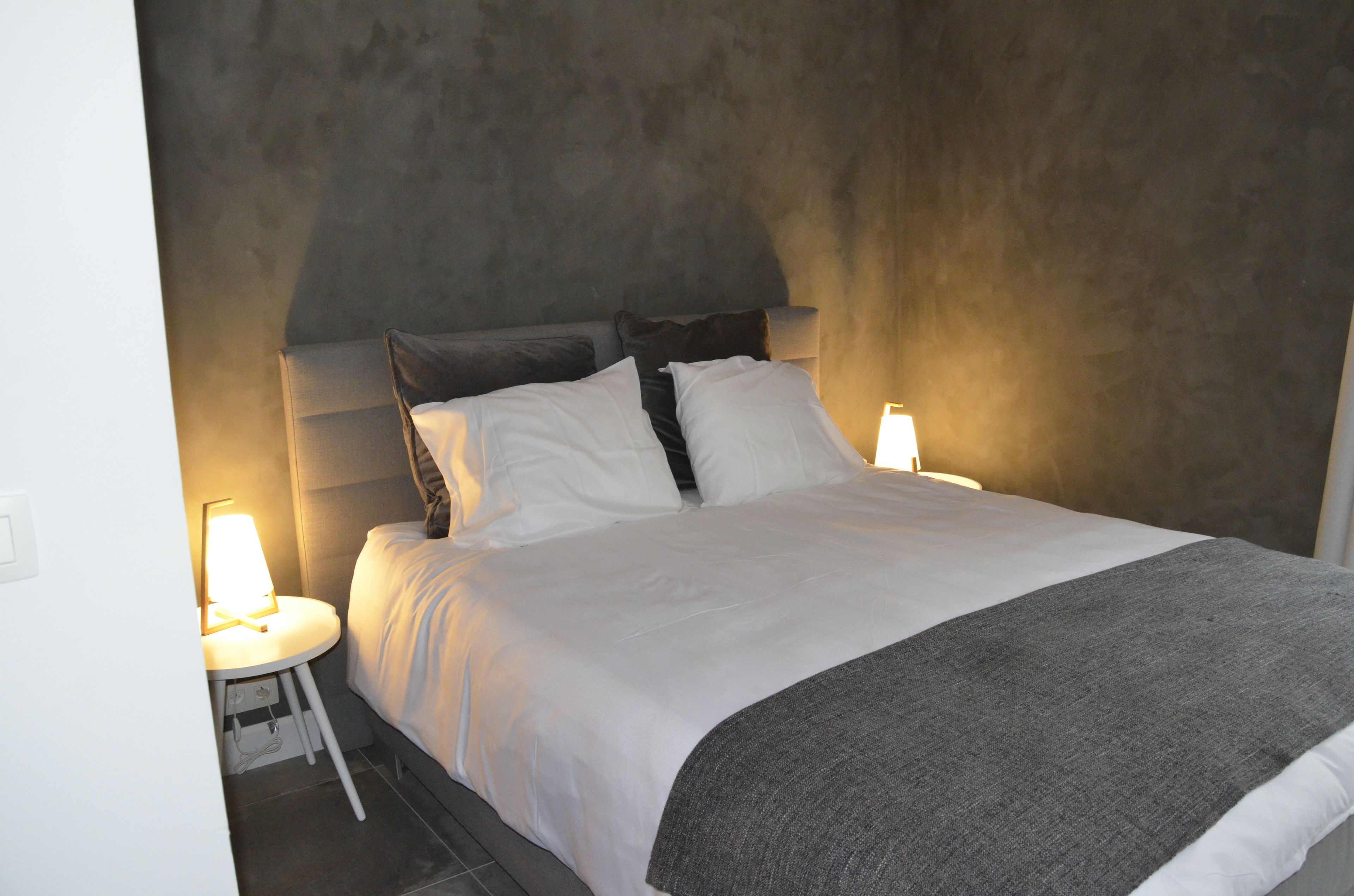 A neatly made bed with white linens and a gray throw blanket, flanked by two bedside lamps.