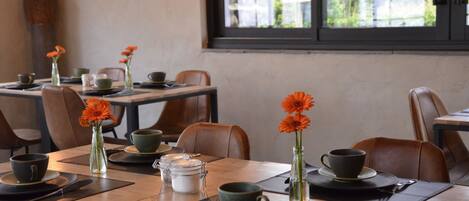 A dining area with tables set for two, each with a cup, saucer, and a vase with orange flowers.