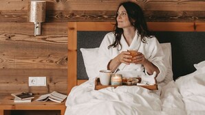 A person sitting on a bed with a tray of breakfast items, a lamp, and a nightstand with books and a power outlet. - Hotel Rosenhof Murau (Murau)