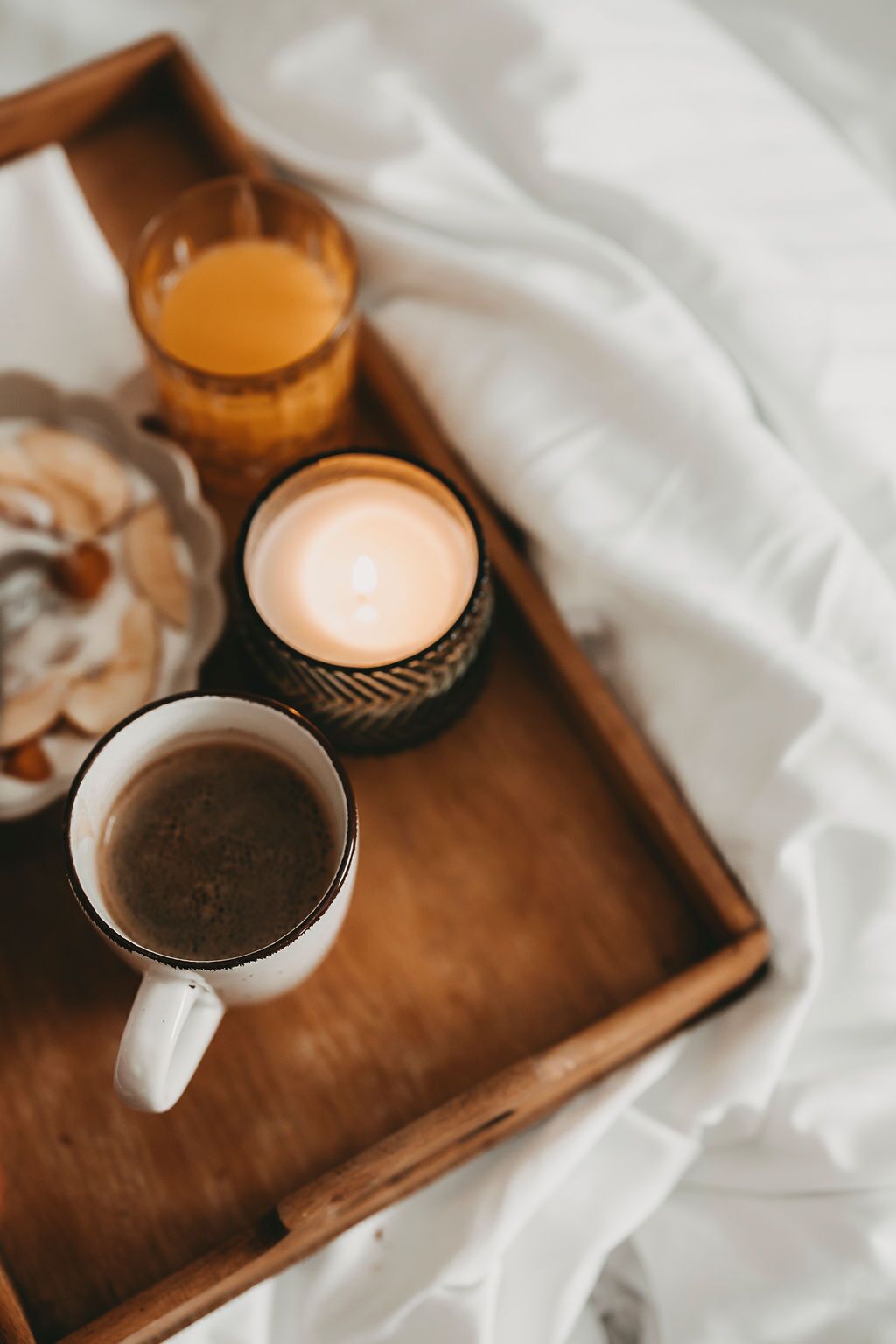 A tray with a white mug of coffee, a lit candle, and a glass of orange juice on a white bedspread.