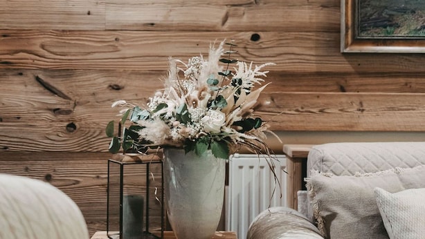 A cozy room with a wooden wall, a framed landscape painting, a white vase with dried flowers, a small table with a candle, and a patterned sofa. - Hotel Rosenhof Murau (Murau)