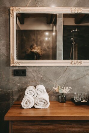 A bathroom with a marble wall, a wooden vanity, a large mirror, and a towel rack with rolled towels.