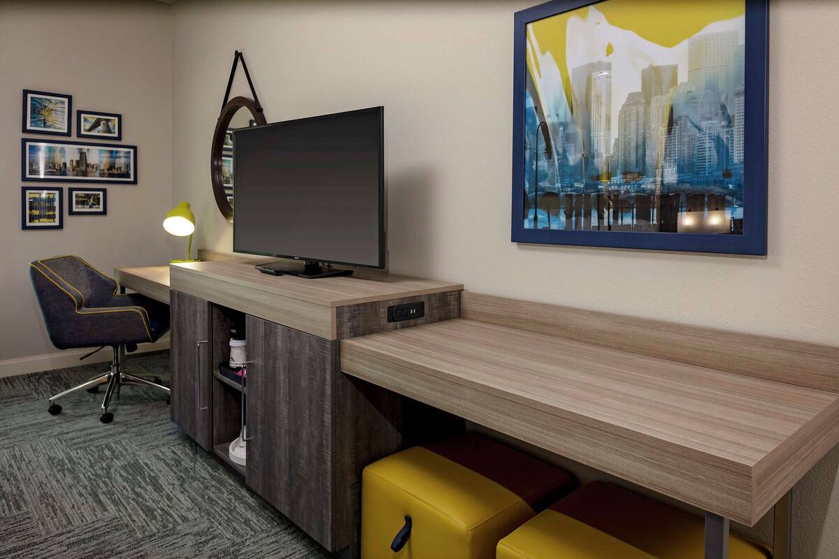 A modern hotel room with a wooden desk, a flat-screen TV, a yellow lamp, and a cityscape painting.