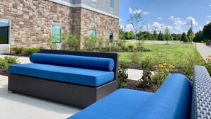 A modern building with stone facade and large windows, outdoor seating area with blue cushions, and a well-maintained lawn with flowers.
