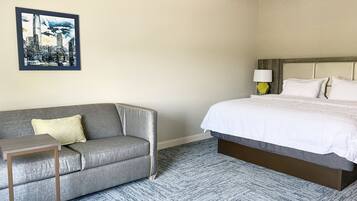 Habitación de hotel con un sofá gris, una cama con ropa blanca, un cuadro de un paisaje urbano en la pared y una lámpara en la mesita de noche.