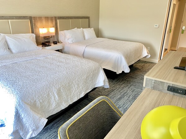 A hotel room with two beds, a desk, a chair, and a lamp.