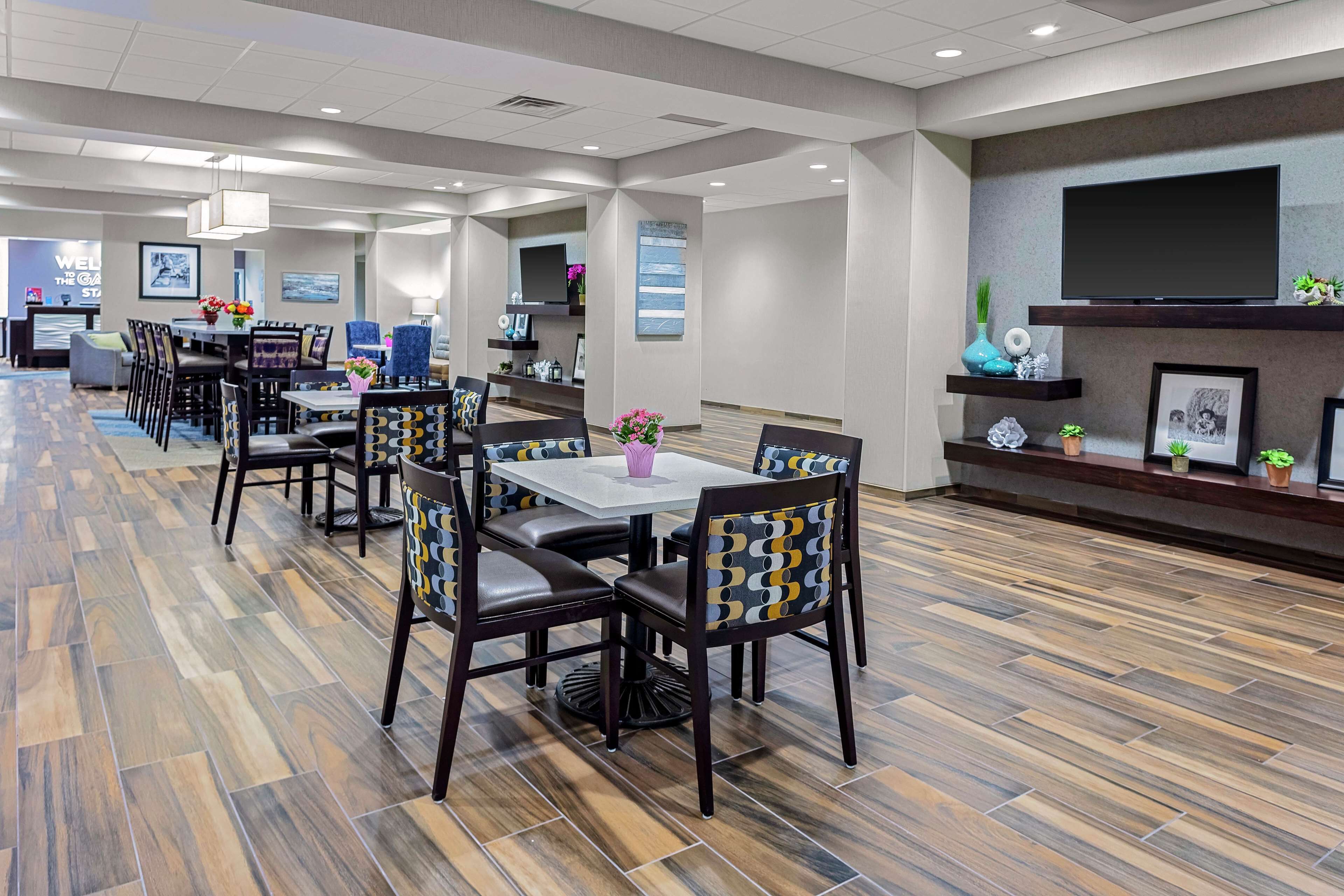 A modern hotel lobby with multiple dining tables and chairs, a flat-screen TV mounted on the wall, and a patterned floor.