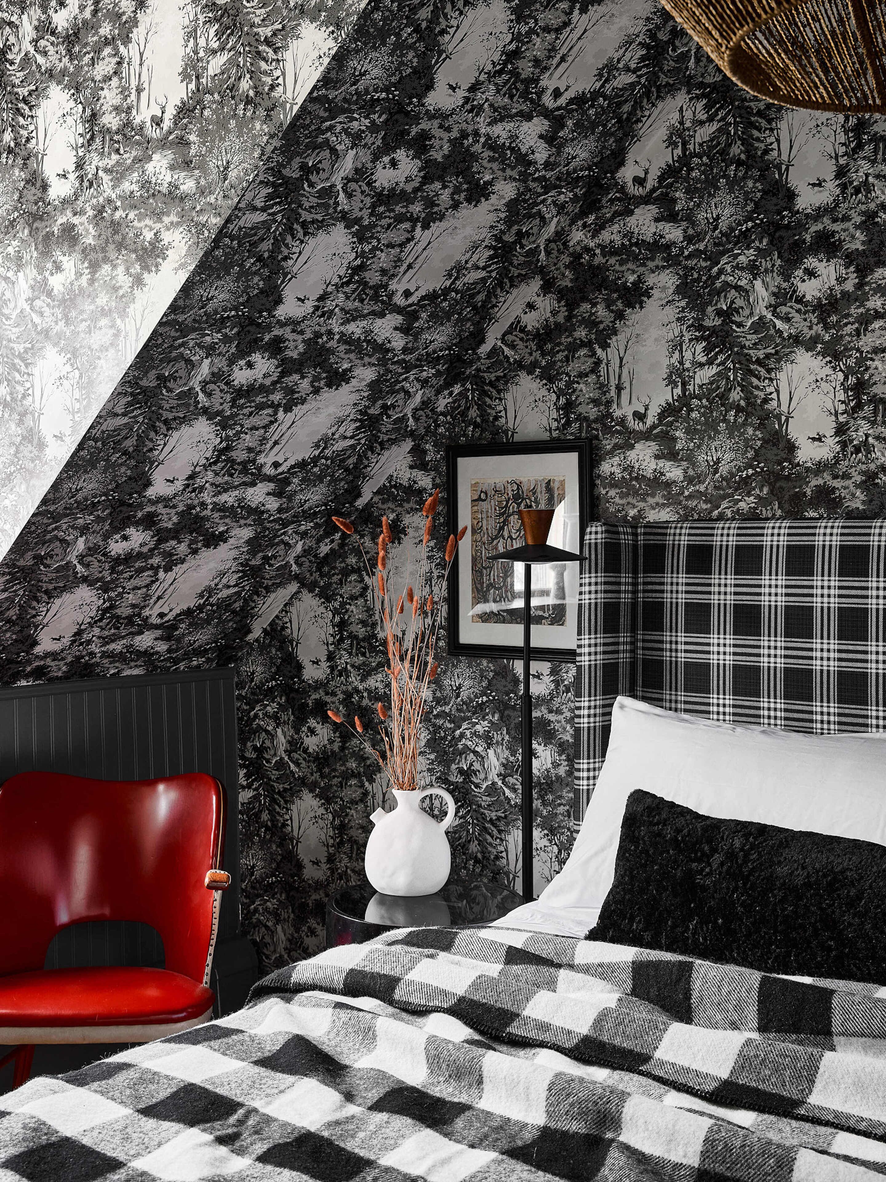 A bedroom with a bed, a red chair, a white vase with plants, a framed picture, and a black and white checkered blanket.