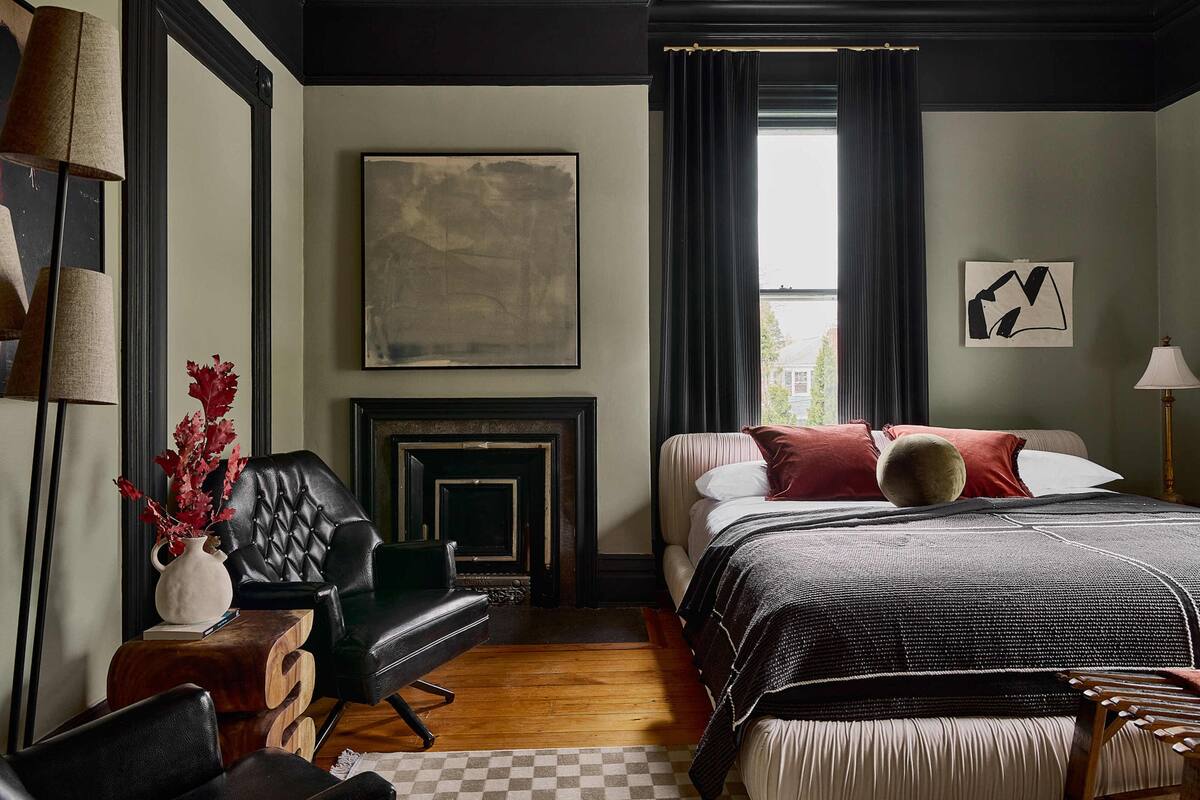 A bedroom with a fireplace, a bed with a black and white bedspread, a black leather chair, and a vase with red flowers.