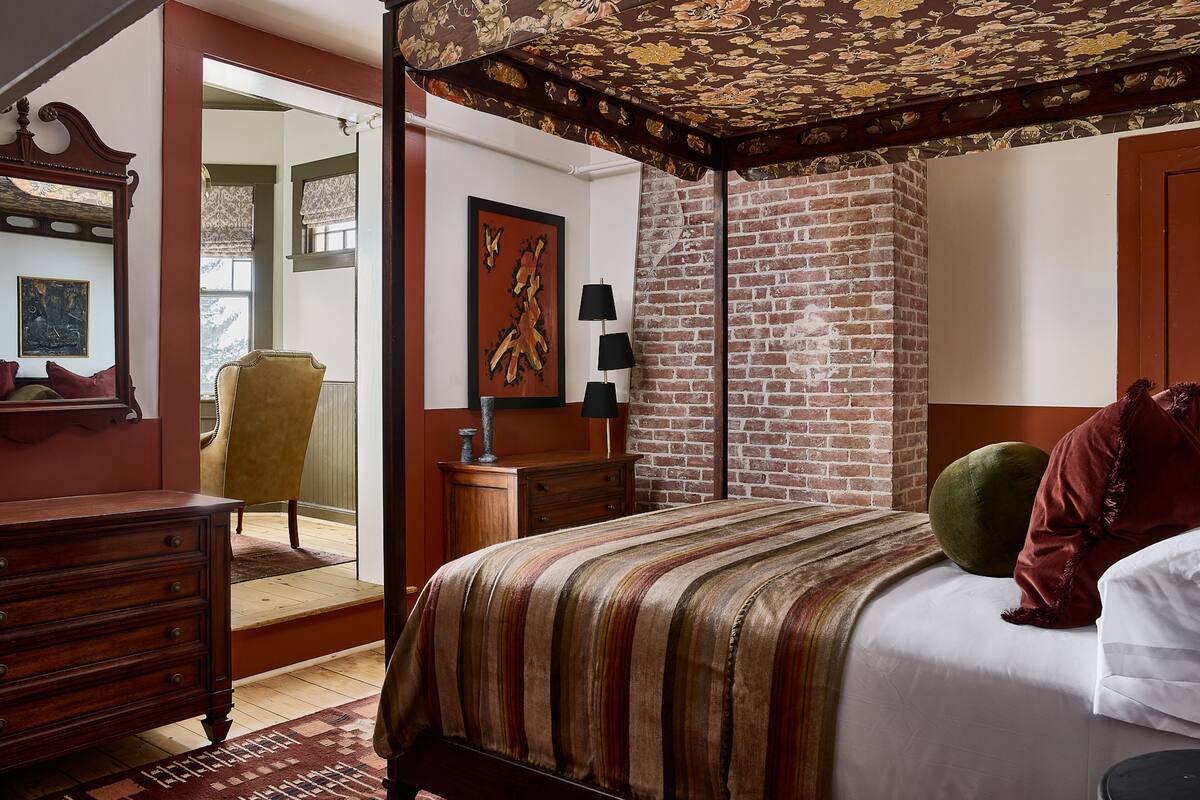 A bedroom with a four-poster bed, a wooden dresser, a chair, and a brick wall.