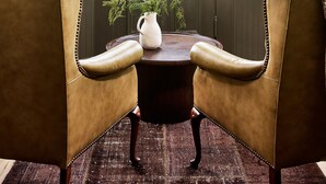A corner room with two leather chairs, a small table, and a view of the outdoors through a large window. - Blind Tiger Burlington (Burlington)