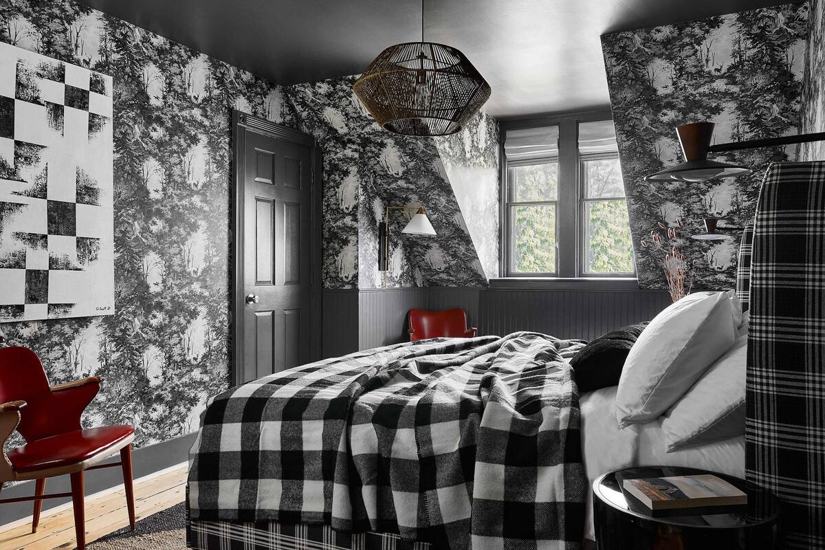 A bedroom with a checkered bedspread, a red chair, a window with a view of trees, and a wall with a forest pattern.