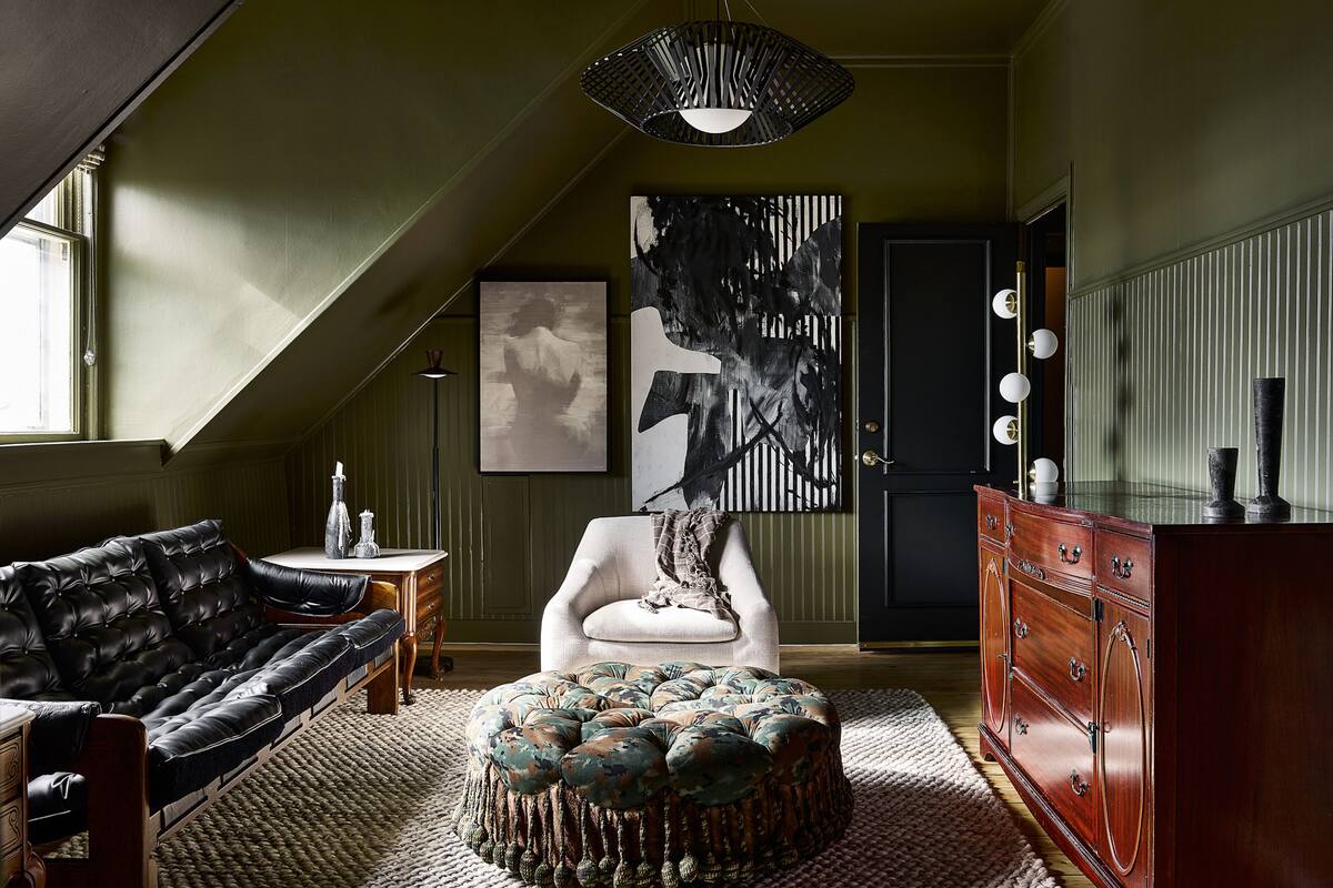 A room with a black leather sofa, a white armchair, a wooden dresser, and a large abstract painting on the wall.