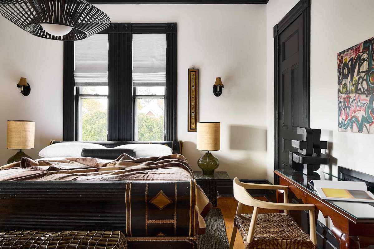A hotel room with a bed, two bedside lamps, a desk with a chair, a window with curtains, and a wall with a framed picture and graffiti art.