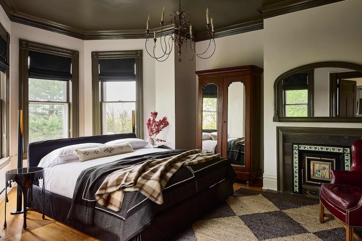A bedroom with a large bed, a fireplace, a mirror, and a window with a view of trees.