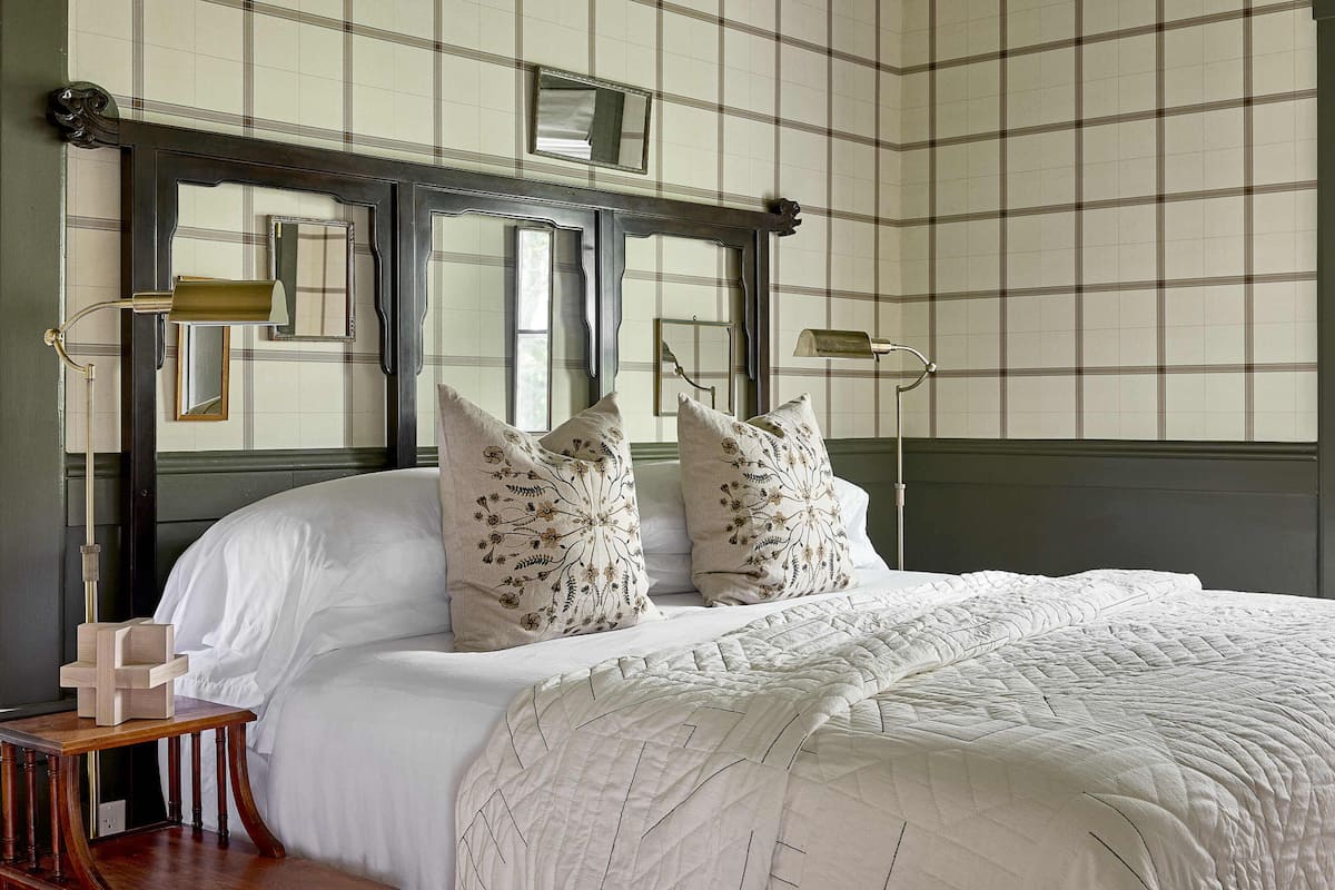A neatly made bed with white linens and floral pillows, a wooden nightstand with a lamp, and a mirror above the bed.
