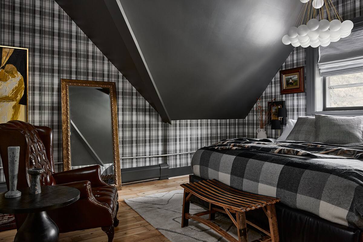 A bedroom with a checkered bedspread, a large mirror, a wooden bench, and a modern chandelier.