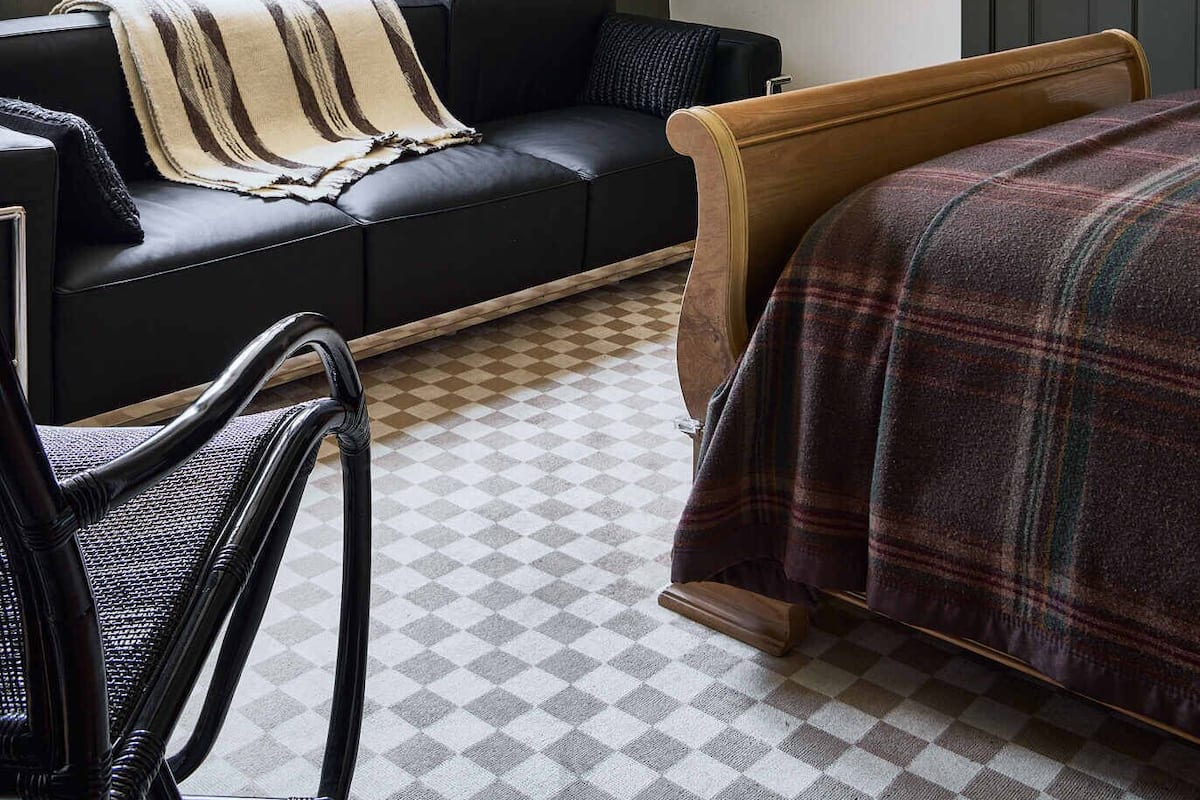 A cozy room with a black leather sofa, a wooden bed, a checkered floor, a large window with blinds, and a woven pendant light.