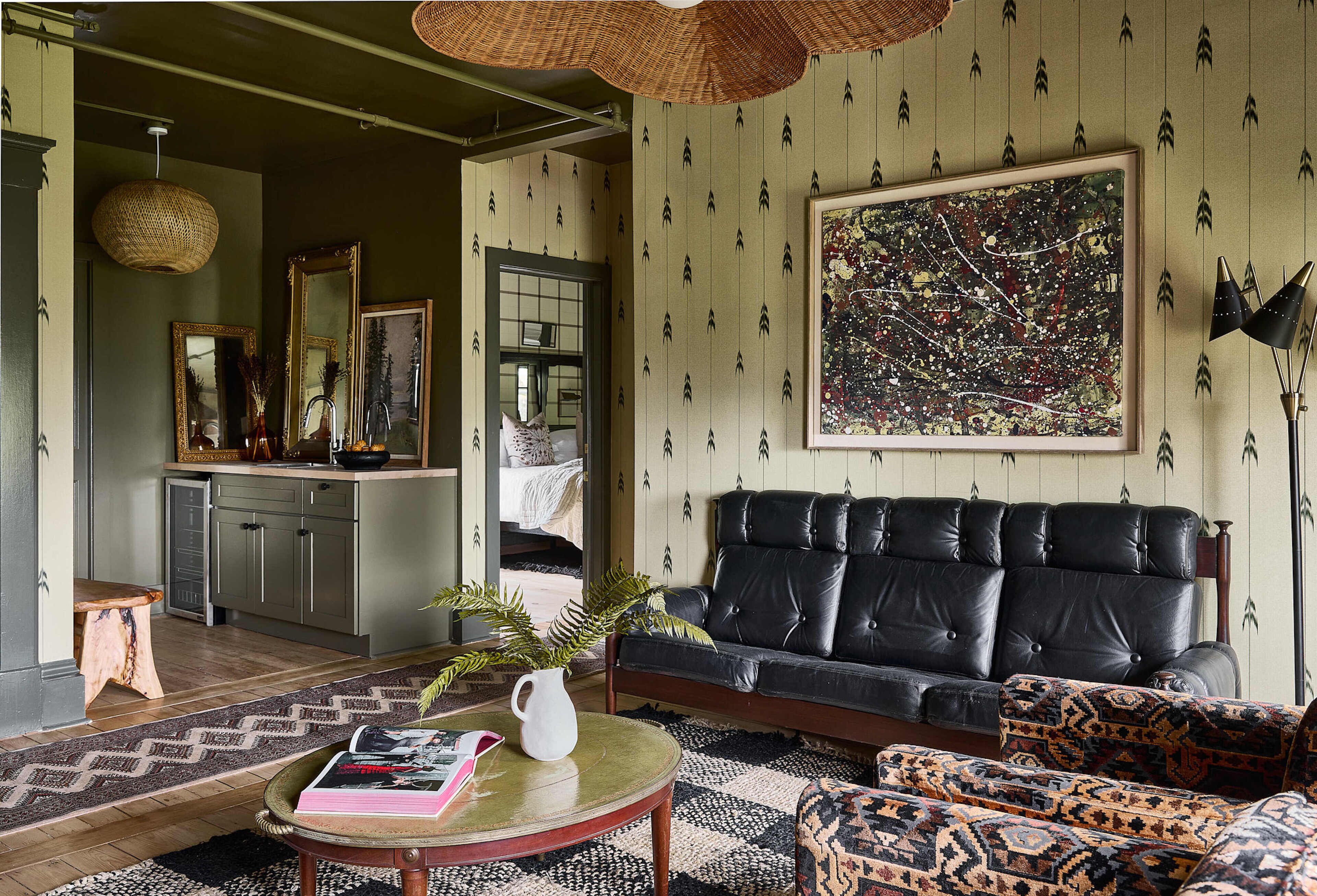 A living room with a black leather sofa, patterned ottoman, and a round coffee table with a book and vase. A large abstract painting hangs on the wall, and a wicker light fixture is above the sofa.