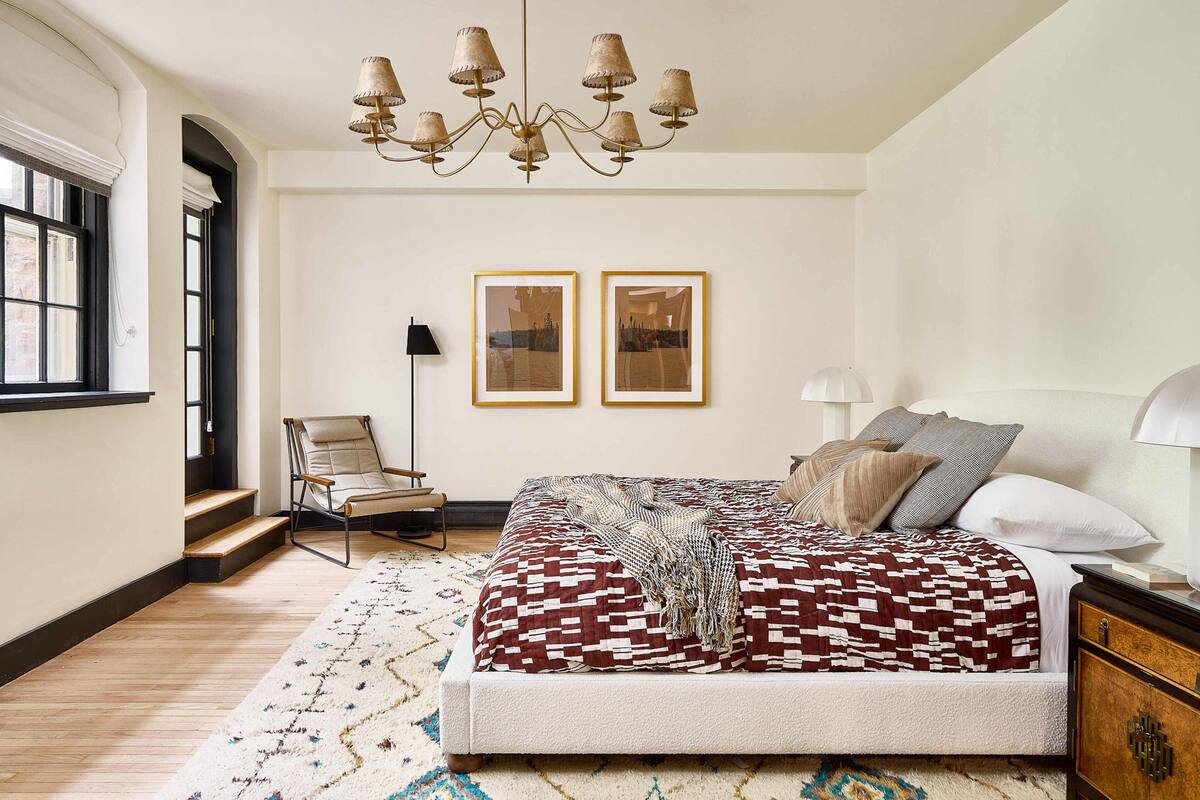 A bedroom with a large bed, a chandelier, two framed pictures on the wall, a chair, and a nightstand.