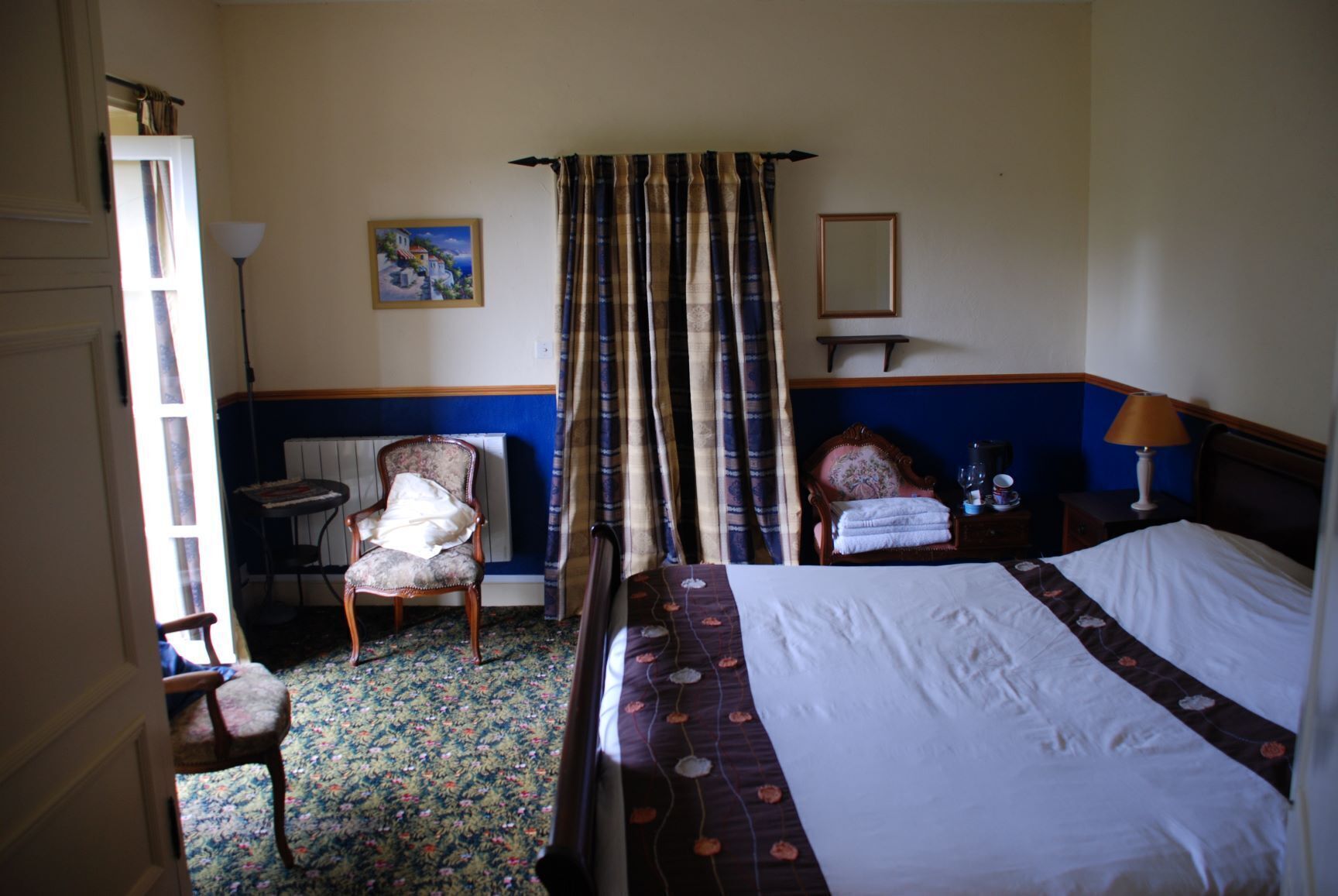 A hotel room with a bed, two chairs, a table, a lamp, and a painting on the wall.