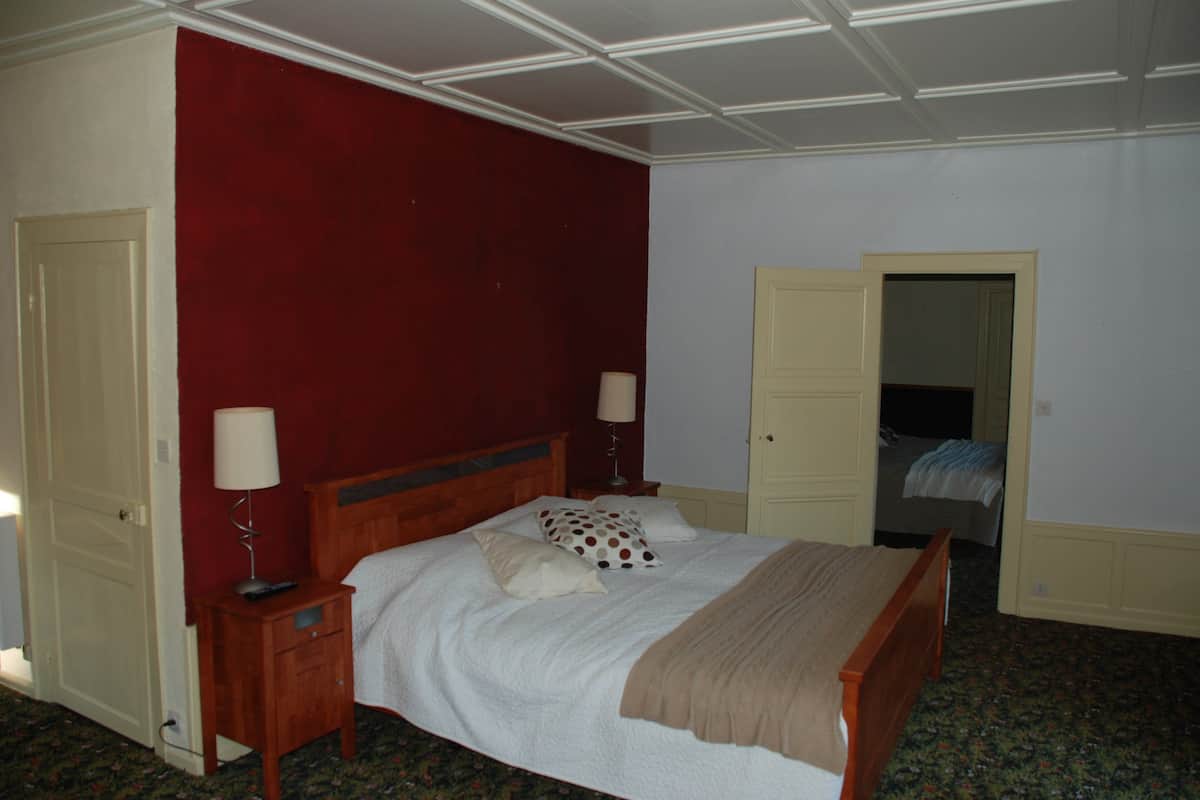 Une chambre à coucher avec un lit en bois, deux tables de chevet avec des lampes, un mur d’accent rouge et une porte donnant sur une autre pièce.