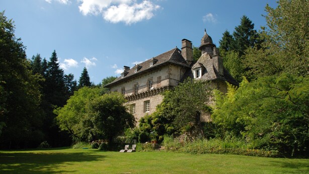 A large, historic stone building with multiple chimneys, surrounded by lush greenery and trees.