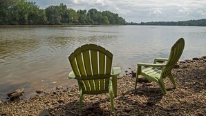 Dos sillas Adirondack verdes sobre una orilla pedregosa junto a un lago con una ribera boscosa al fondo.