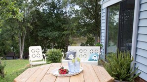 Mesa de madera en un patio con una bandeja de fresas y bebidas, rodeada de sillas blancas con cojines estampados, frente a un fondo de follaje y una casa.