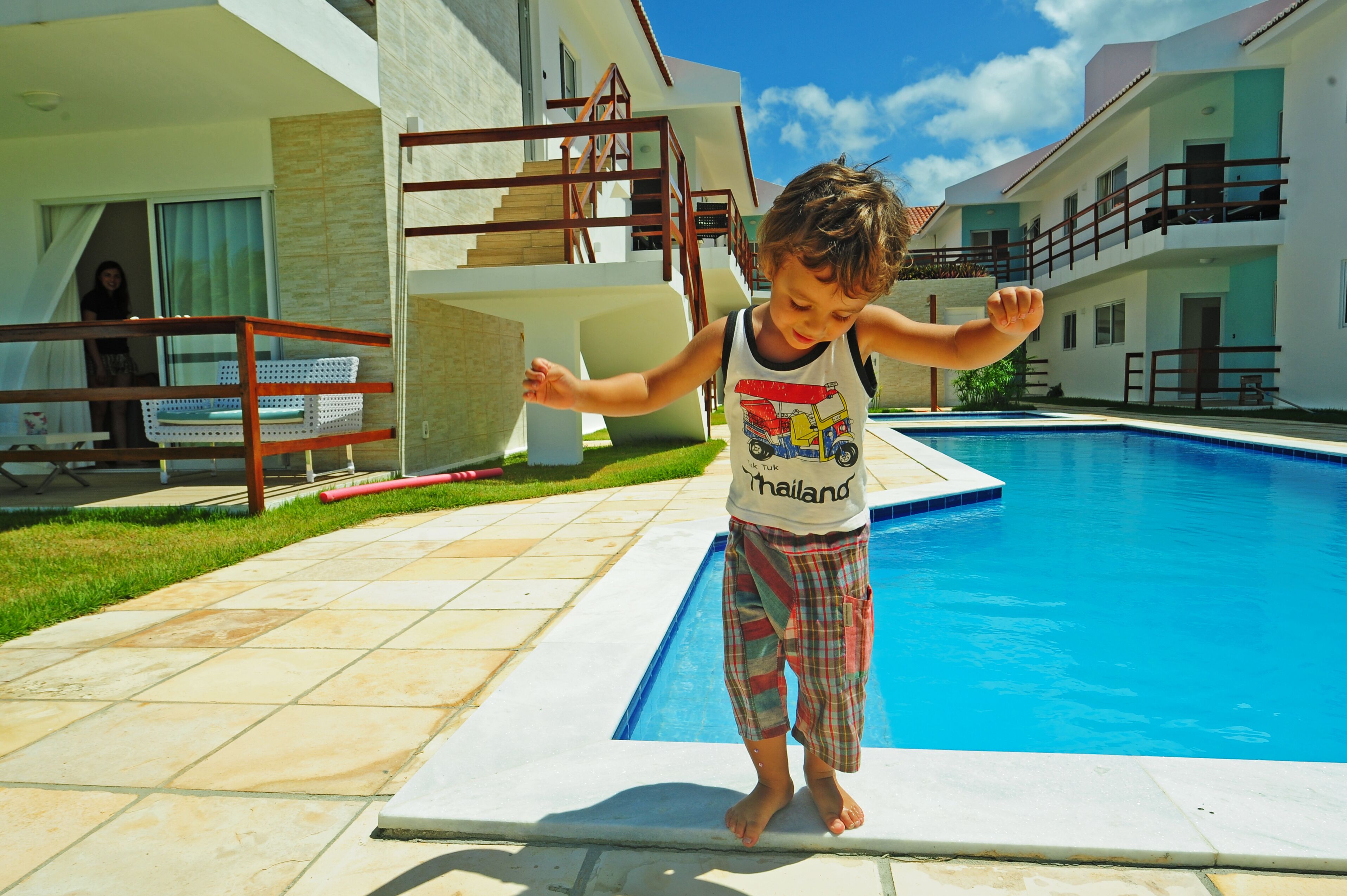 Ein Kind steht auf einer Pool-Plattform mit einem klaren, blauen Pool, einer Holzterrasse und einem Gebäude mit Balkon im Hintergrund.