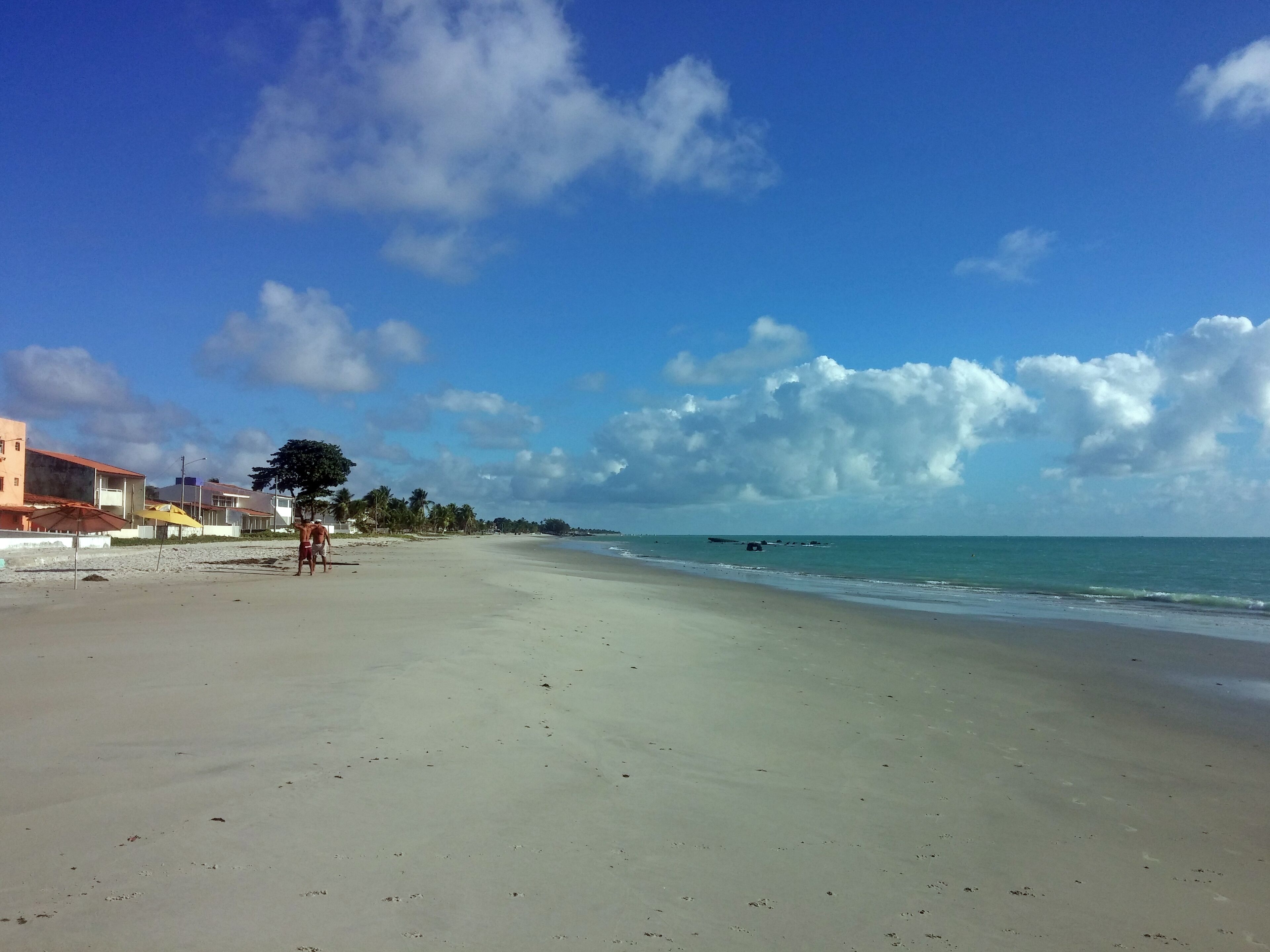 Une plage avec un ciel dégagé, une mer calme et quelques bâtiments en arrière-plan.