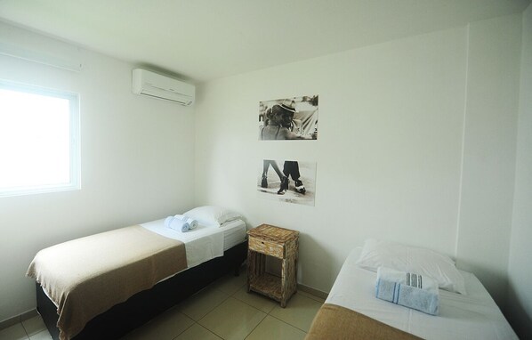 A small, clean room with two single beds, a small wooden side table, and a window providing natural light.