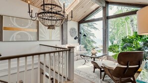 Una habitación amplia con techo de madera, un ventanal grande, una araña y vistas al verdor exterior.