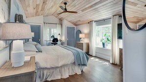 Un dormitorio con techo de madera, una cama con colcha azul, dos lámparas de noche, un escritorio con silla y una ventana con cortinas.