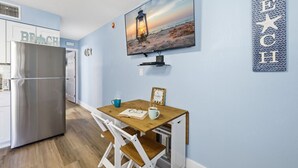 A modern kitchen with a stainless steel refrigerator, a wooden dining table with chairs, a TV mounted on the wall, and a beach-themed decor.