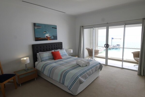 Un dormitorio con cama, mesita de noche, silla, ventana con vista al mar y un cuadro en la pared.