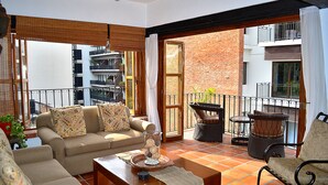 Un salon comprenant un canapé, une table basse et des fauteuils, un balcon donnant sur un immeuble en briques, et une fenêtre offrant une vue sur un balcon.