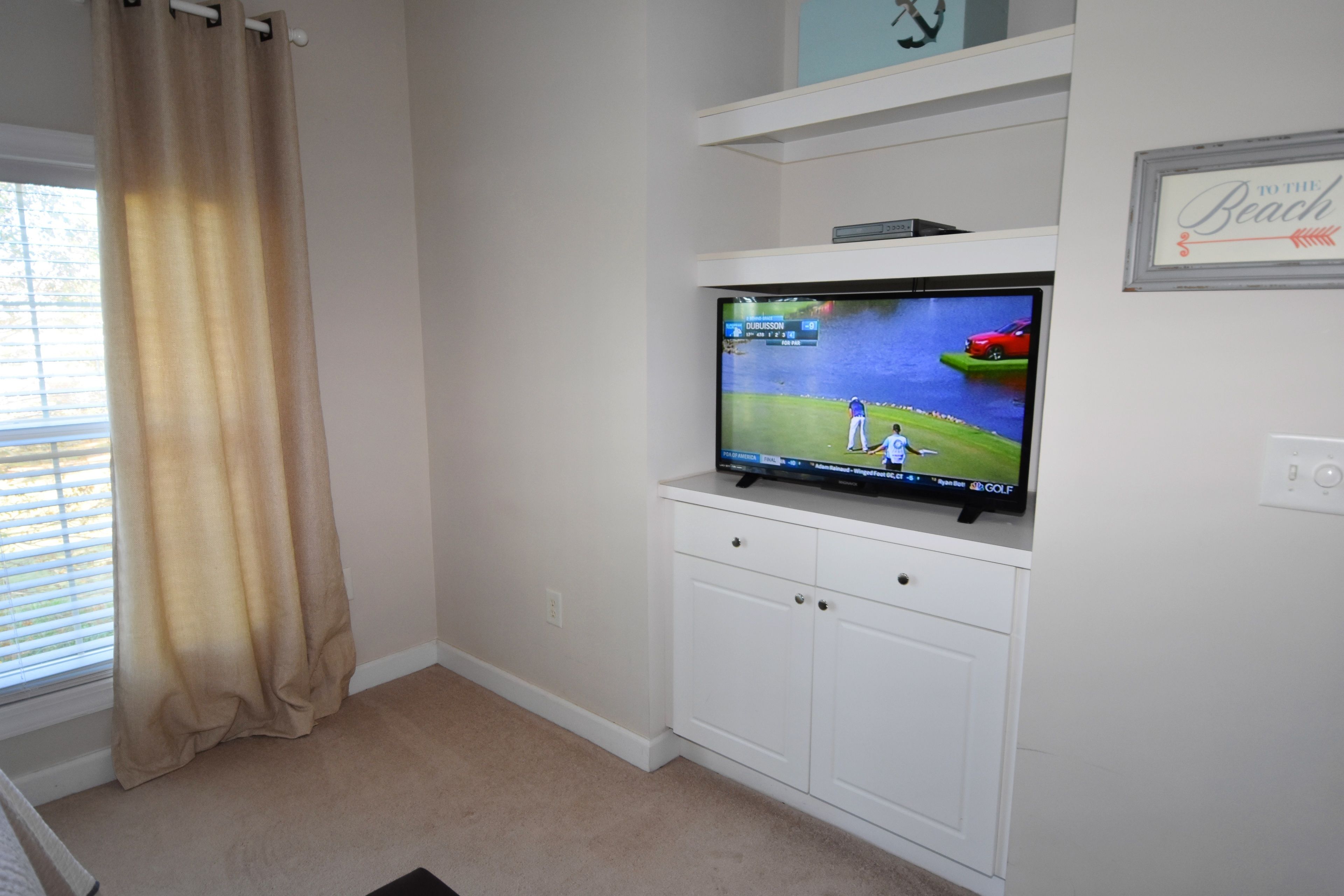 Una habitación con una televisión montada en un mueble blanco, una ventana con cortinas color beige y un letrero en la pared que dice "TO THE BEACH".