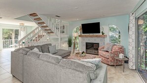 Amplia sala de estar con un sofá seccional gris, un sillón estampado, una chimenea y una escalera que lleva a un balcón.